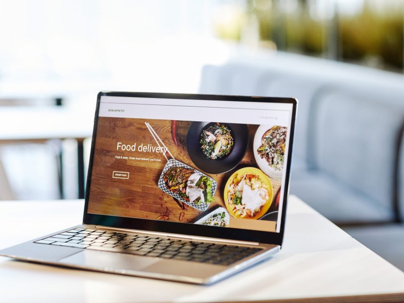 Background image of opened laptop with Food delivery website on table in food court or cafe interior, copy space
