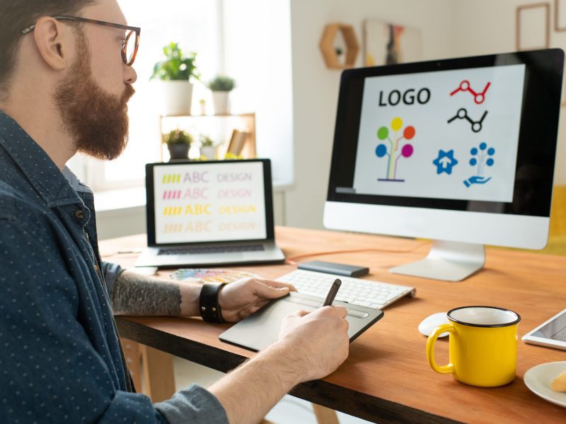 Over shoulder view of concentrated bearded designer in glasses sitting at desk and using drawing pad while working on website graphics